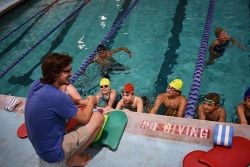 A Barracudas coach crouches at the edge of the pool with seven swimmers looking up at him.